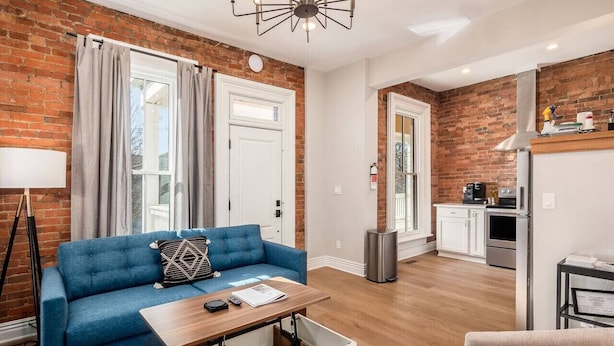 A modern apartment with a blue sofa, a wooden coffee table, a kitchen area with appliances, and a brick wall. - Access OSU & Crew Stadium | Pet Friendly | Parking (Columbus)