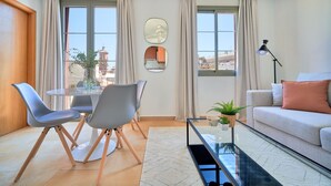 A modern living room with a round table, two grey chairs, a sofa with a cushion, a floor lamp, and a view of buildings through large windows.