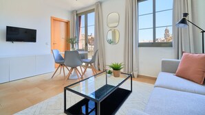 A modern living room with a glass coffee table, a sofa, a TV mounted on the wall, and a dining area with chairs and a table.