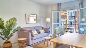 Ein modernes Wohnzimmer mit einer Couch, einem Esstisch und Stühlen. Eine Topfpflanze und ein gerahmtes Bild sind an der Wand zu sehen.