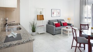 Un salon moderne avec un canapé gris, des coussins rouges, une table basse blanche et un coin repas comprenant une table en verre et des chaises à rayures.