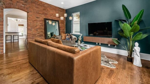 A modern living room with a brick accent wall, a large sofa, a flat-screen TV, and a large green plant. - Private Patio |German Village| Walk Everywhere (Columbus)