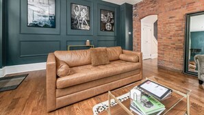 A living room with a brown leather sofa, a glass coffee table, and framed artwork on the wall. - Private Patio |German Village| Walk Everywhere (Columbus)