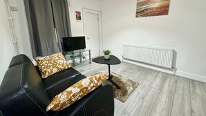 A modern living room with a black sofa, a glass coffee table, a TV on a stand, a radiator, and a painting on the wall.