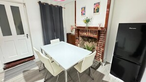 A dining area with a white table and chairs, a black refrigerator, a brick fireplace, and two framed floral artworks on the wall.