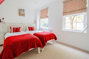 Two single beds with white headboards and red bedding, a large window with a view of trees, and a small painting adorn this cozy space.