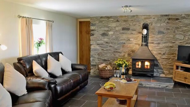 A cozy living room with a stone wall, a leather sofa, a wooden coffee table, and a fireplace. - Golygfar Mynydd Retreat - Sleeps 6 (Tregaron)