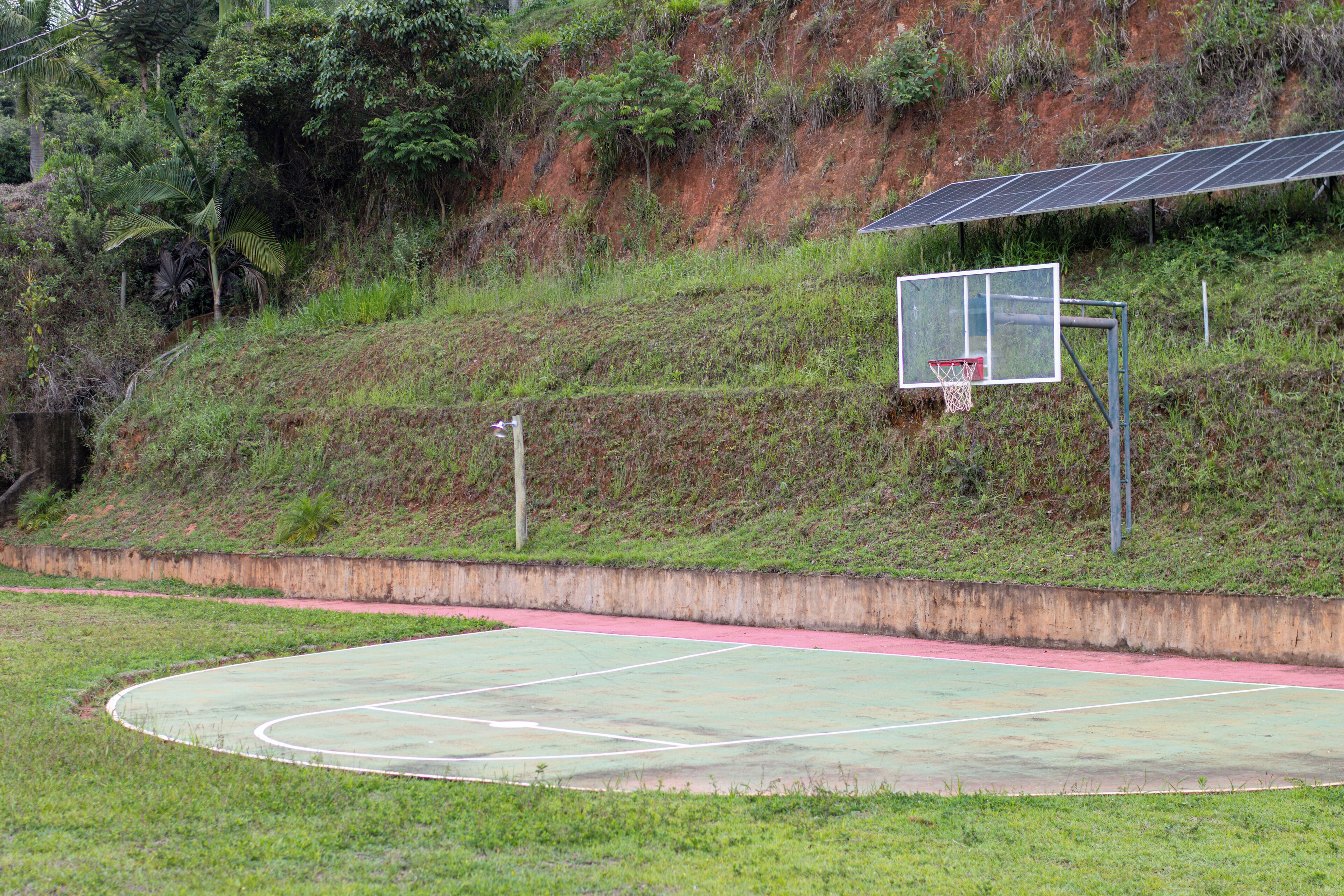 An outdoor basketball court with a red track, surrounded by greenery and a hillside.