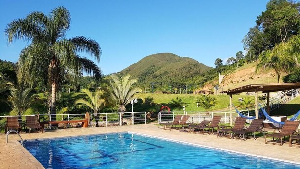 A swimming pool with lounge chairs and palm trees, surrounded by a hilly landscape. - Pousada Nefelibatas (Águas de Lindóia)