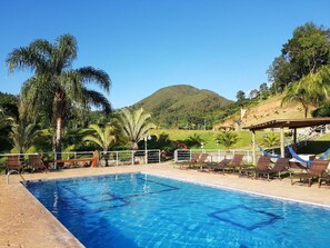 A swimming pool with lounge chairs and palm trees, surrounded by a hilly landscape. - Pousada Nefelibatas (Águas de Lindóia)
