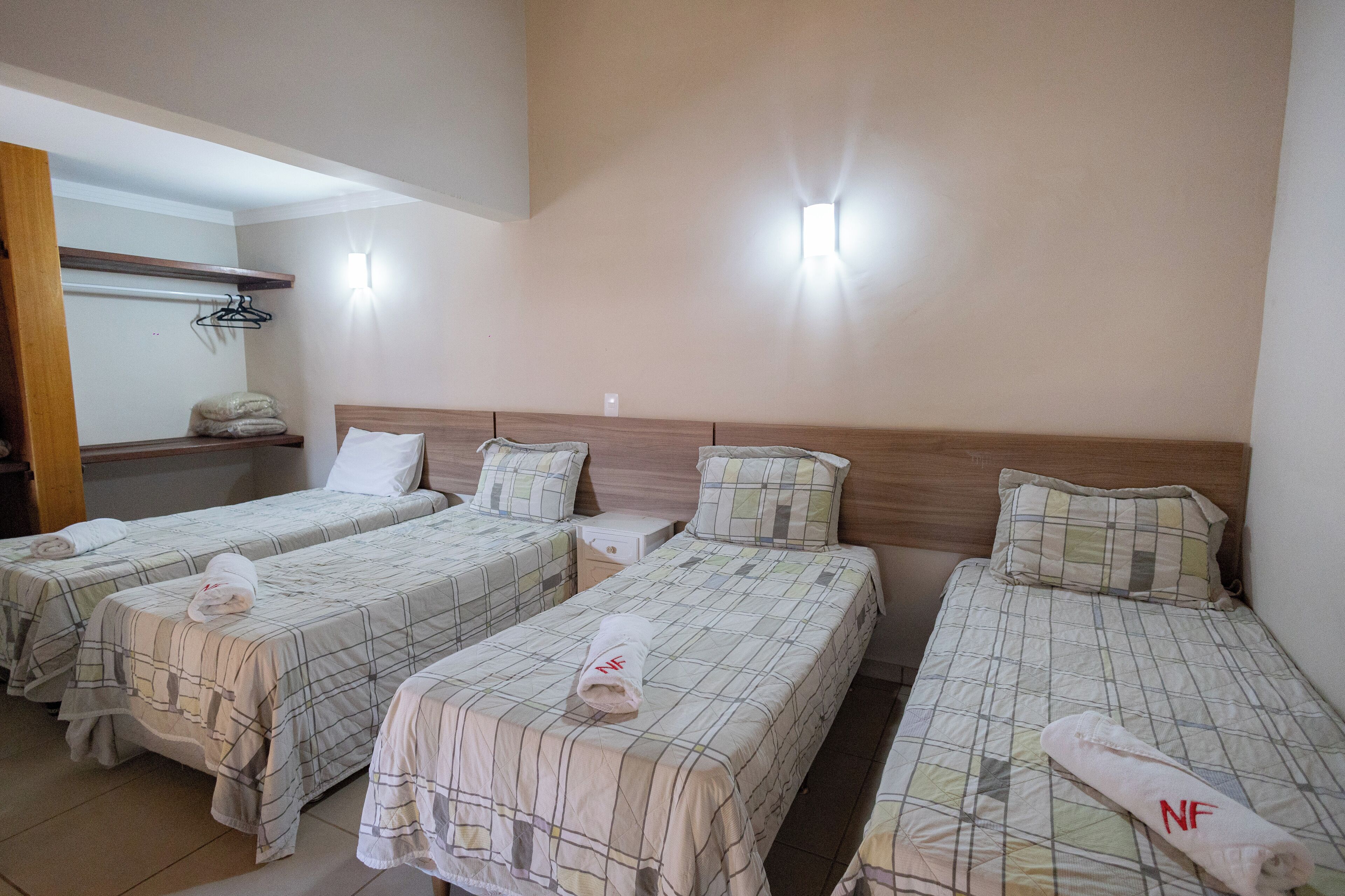 A hotel room with three single beds, each with a neatly folded towel and a pillow. There is a wooden headboard, a small bedside table, and a wall-mounted light fixture.
