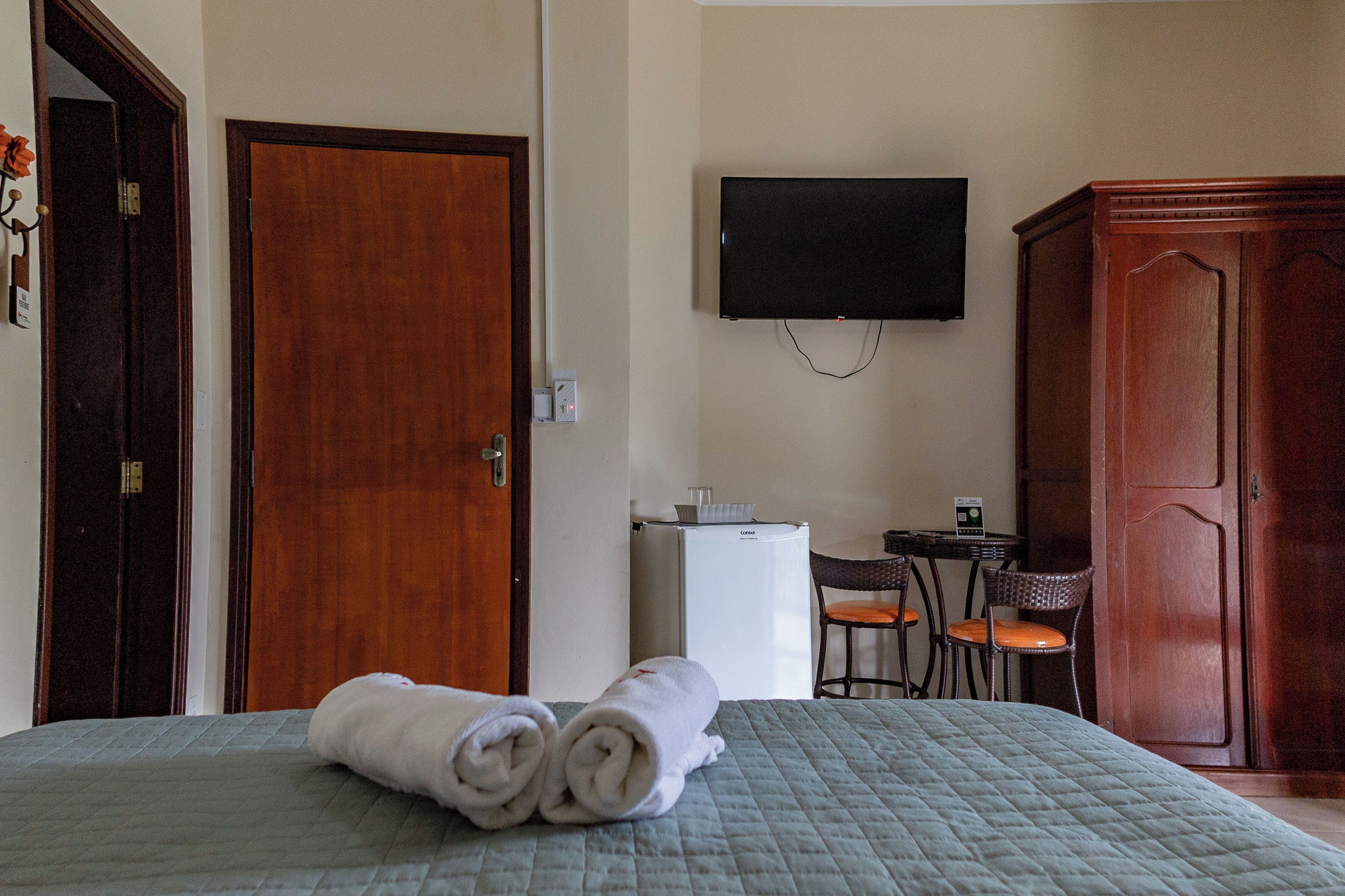 Quarto de hotel com cama, televisão na parede, guarda-roupa de madeira, mesa pequena com cadeiras e frigorífico.