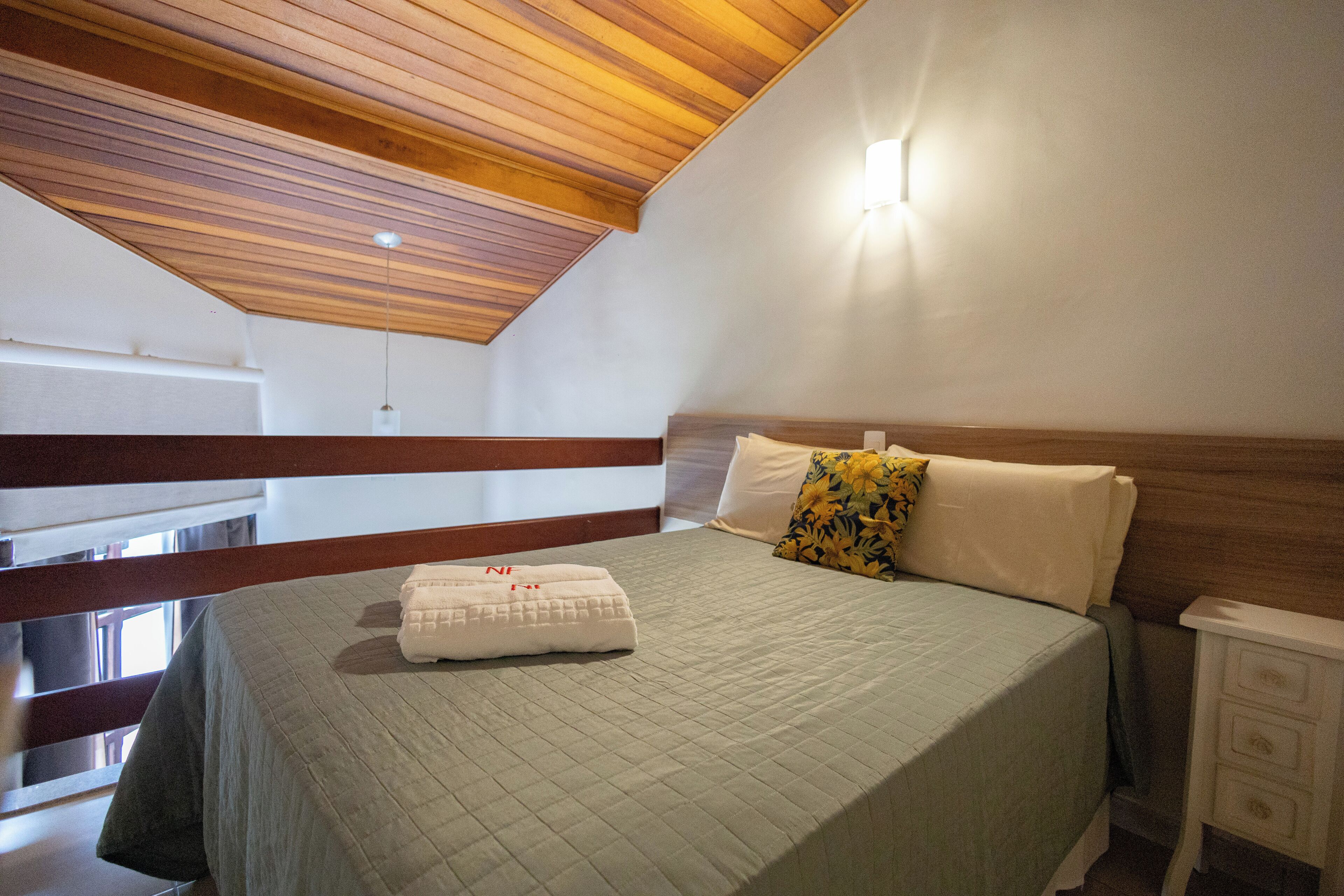 Un dormitorio con techo de madera, una cama con colcha, un almohadón y una mesita de noche.