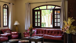 A room with a red leather sofa, a wooden coffee table, wicker side tables, and a large arched window with curtains. - Pousada Nefelibatas (Águas de Lindóia)