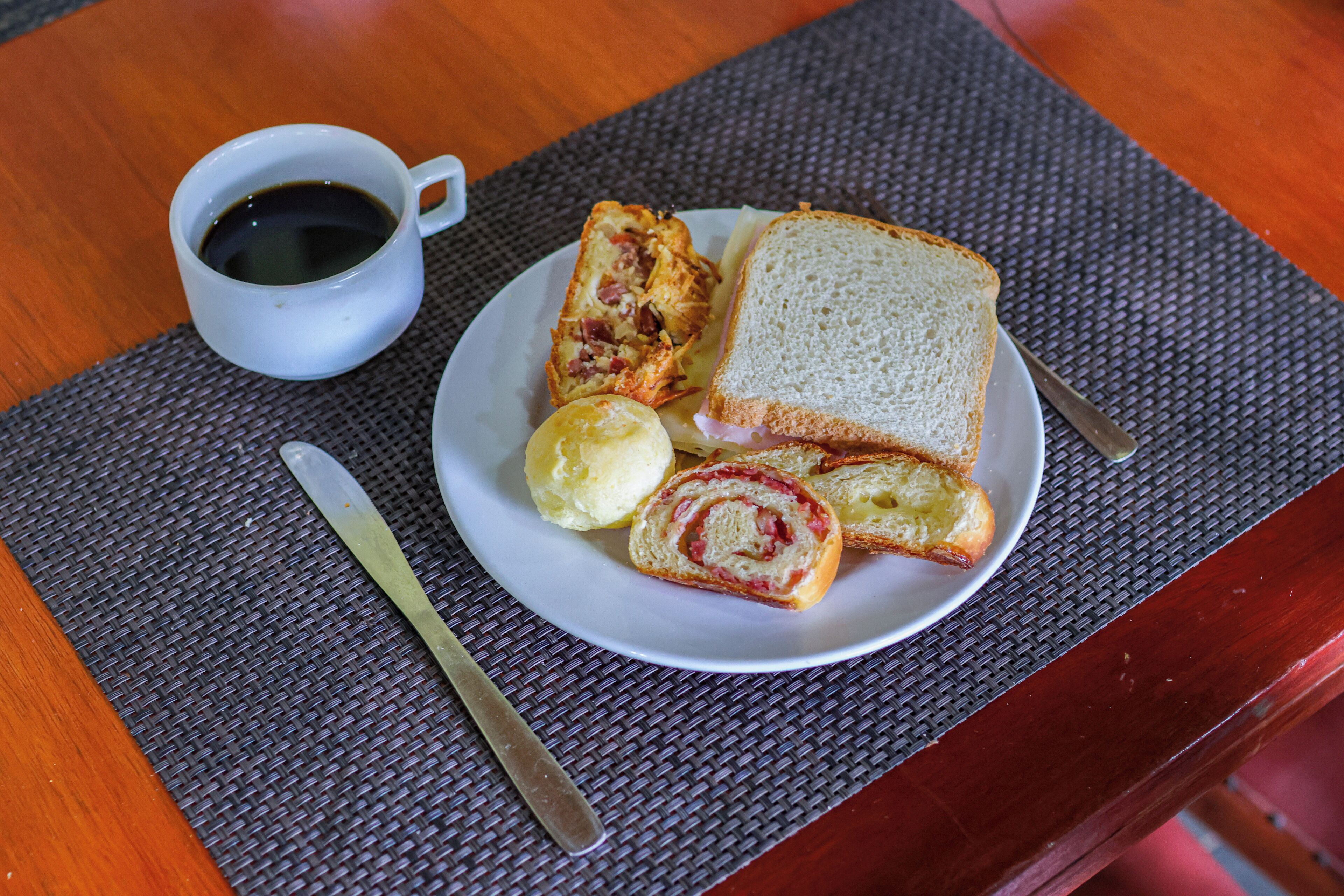 Ein Teller mit einem Sandwich, einer Backware und einer Kaffeetasse auf einem Tisch mit Unterlage.