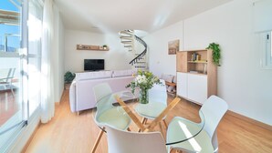 A modern living room with a glass dining table, white chairs, a sofa, a TV, a spiral staircase, and a bookshelf.