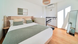 A modern bedroom with a large bed, a wooden headboard, a bedside table, a hanging light fixture, a mirror, and a staircase.