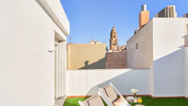 A rooftop terrace with two sun loungers, a small table with a drink and flowers, and a view of a historic tower in the background.