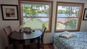 Une pièce comprenant un lit, une table à manger avec des chaises et deux fenêtres donnant sur un lac et des bâtiments.