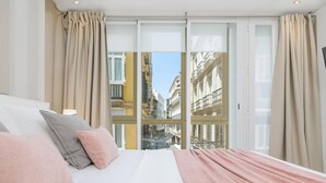 A hotel room with a large bed, beige curtains, and a view of a cityscape through floor-to-ceiling windows.