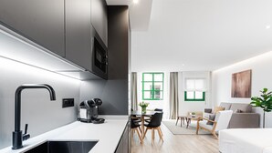 A modern kitchen with a built-in stove, a coffee machine, and a sink. There is a dining area with a table and chairs, and a living room with a sofa and a plant. - Barroso II - iloftmalaga (Málaga)