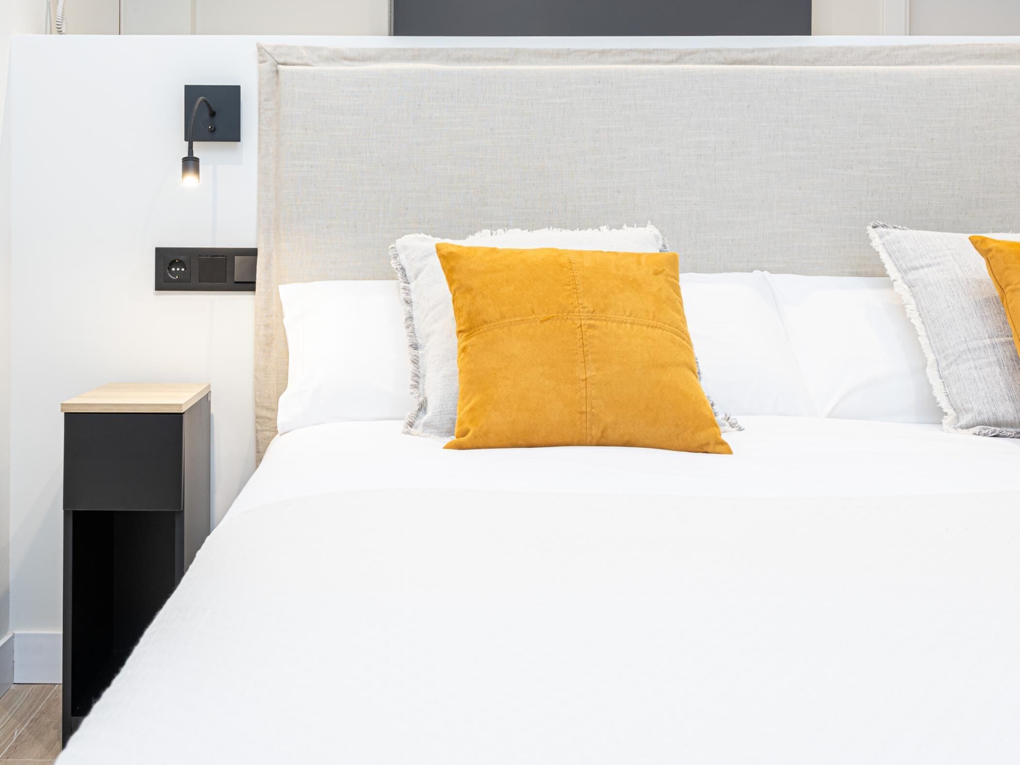 Quarto de hotel moderno com uma cama grande, uma mesinha de cabeceira e um painel de controle fixado na parede.