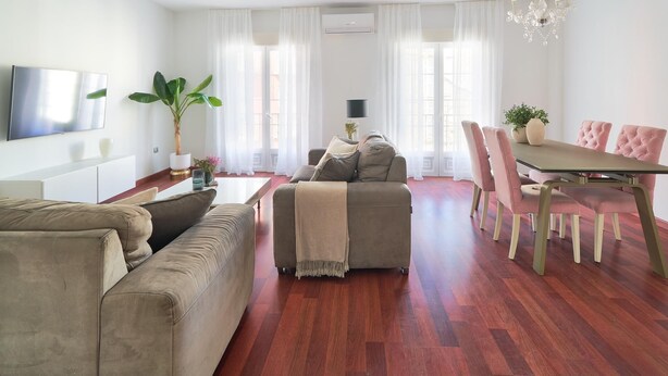 Una sala moderna con sofá, reposapiés y una mesa de comedor con sillas. Hay un televisor montado en la pared, una planta y una araña.