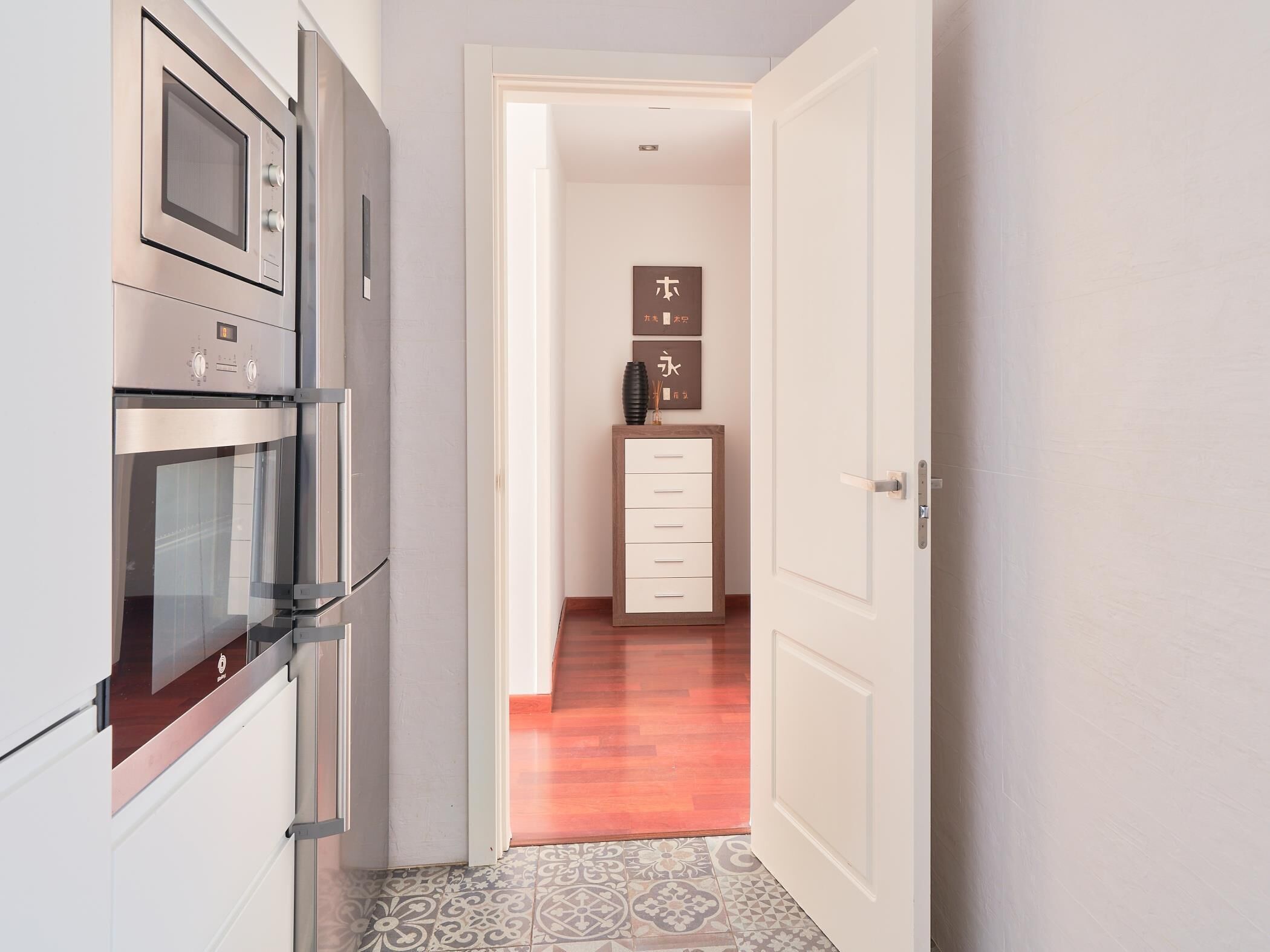 A narrow hallway with a modern kitchen on one side, featuring stainless steel appliances. A wooden cabinet and a framed artwork are visible in the background.