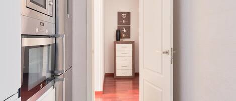 A narrow hallway with a modern kitchen on one side, featuring stainless steel appliances. A wooden cabinet and a framed artwork are visible in the background.