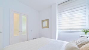 Un dormitorio con una cama grande, una mesita de noche con un florero, una ventana con persianas y una puerta con paneles de vidrio esmerilado.