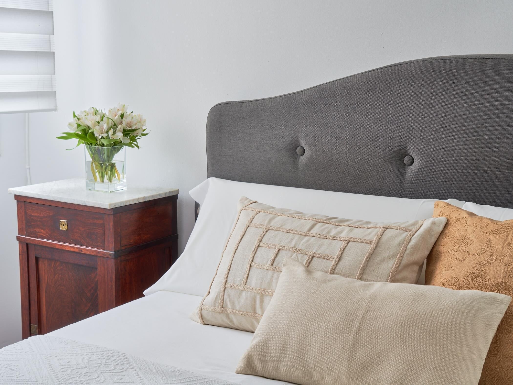 Una cama bien tendida con un cabecero tapizado en gris, una mesita de noche de madera con un florero, y una pared blanca.
