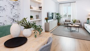 A modern living room with a sofa, coffee table, and a dining area with a table and chairs. There is a bookshelf with decorative items and a TV mounted on the wall.