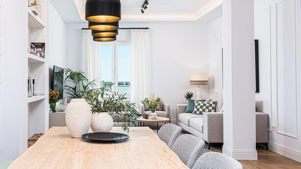 A modern living room with a wooden dining table, a sofa, and a coffee table. There is a large window with curtains, a bookshelf, and a hanging pendant light.