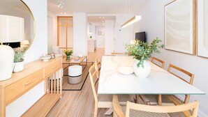 Un comedor moderno con una mesa de vidrio, sillas de madera y juego de comedor blanco. Hay un espejo redondo sobre una consola de madera, un florero con flores y un televisor en la pared.