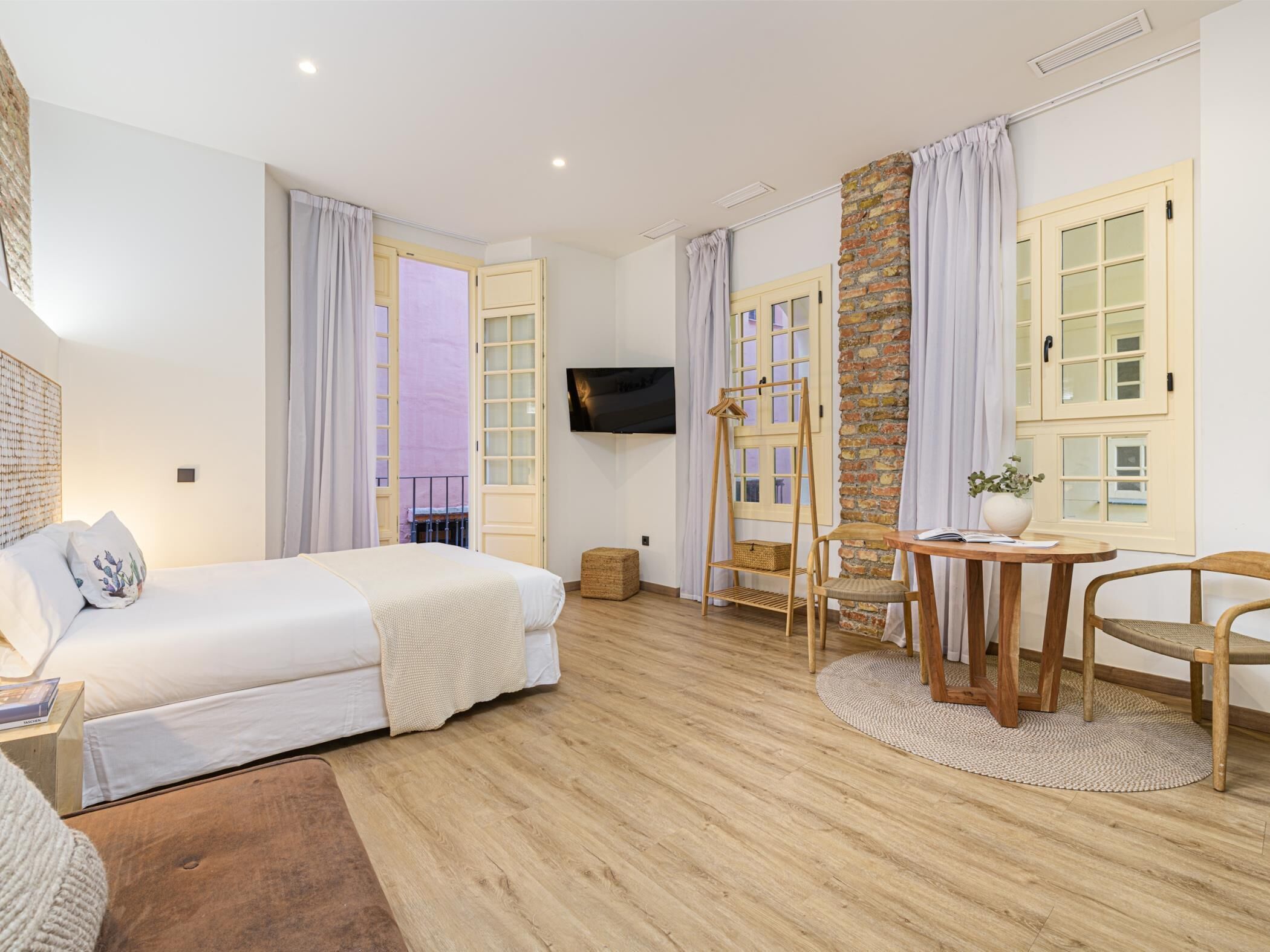A hotel room with a bed, a TV, a wooden table with chairs, and a stone wall.