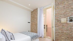 A bedroom with a brick wall, a bed with white bedding, a wooden floor, and a view of a balcony through an open door.