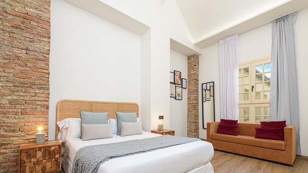A bedroom with a large bed, a wooden nightstand, a stone wall, a stone pillar, a wooden floor, a window with curtains, and a red sofa.