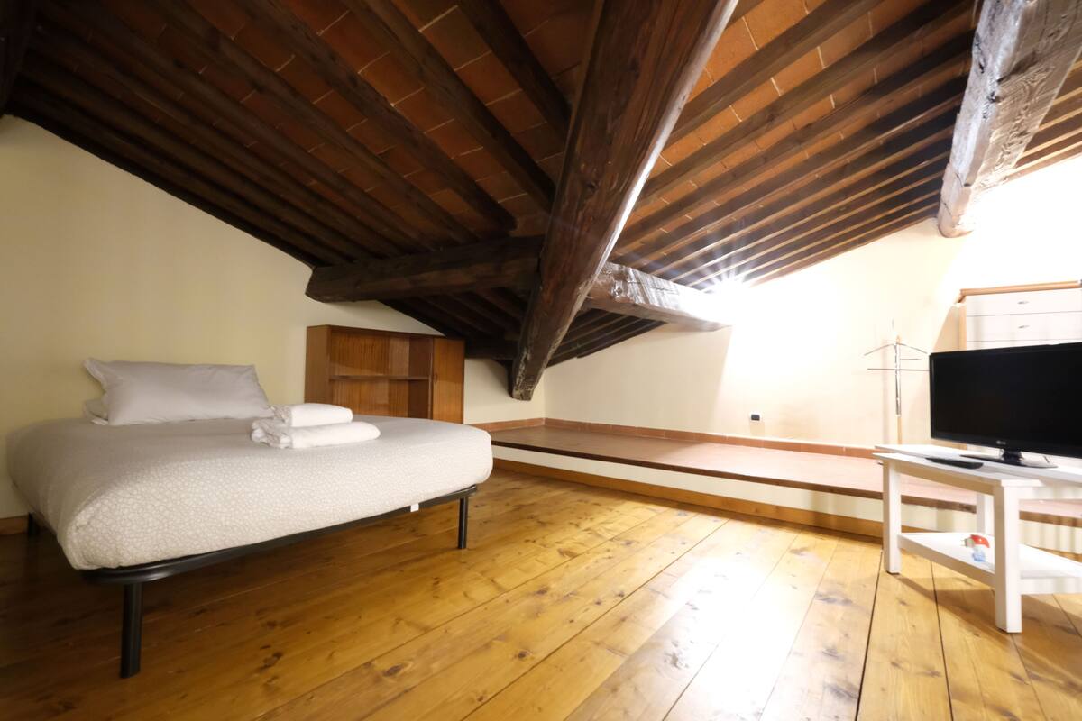Een slaapkamer met een houten plafond, een bed met een witte matras en kussens, een houten vloer, een tv op een witte standaard, en een raam met natuurlijk licht.