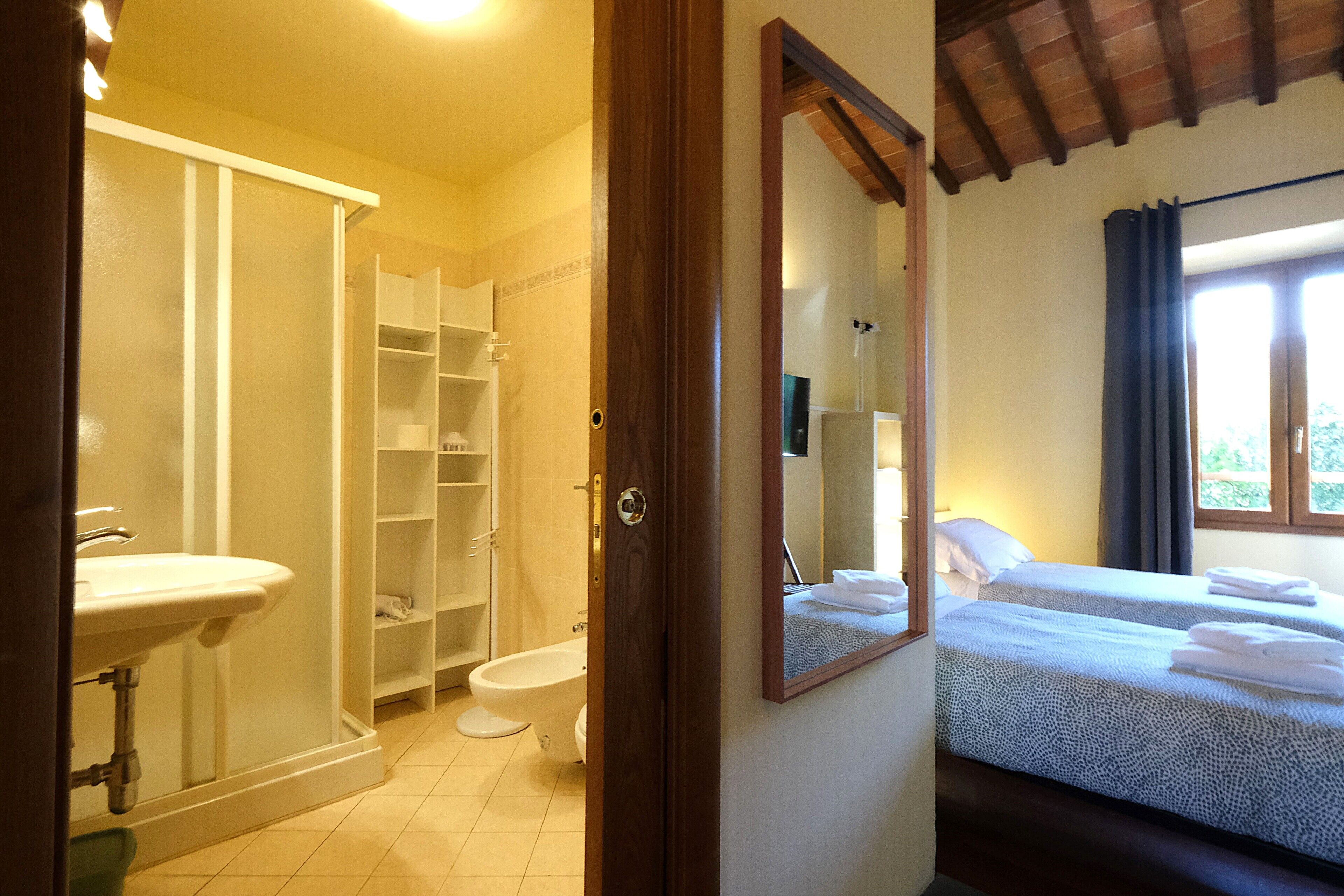 A bathroom with a sink, toilet, and shower. There is a bed with a mirror and a window with curtains.