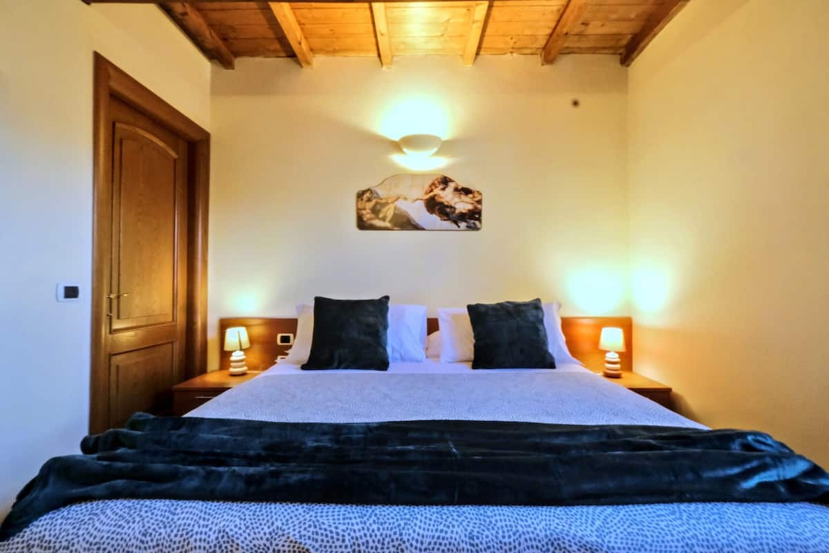 Een slaapkamer met een houten plafond, een bed met een zwarte deken, wit beddengoed, twee bedlampen en een wandlamp.