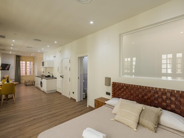 Habitación de hotel moderna con una cama grande, cabecera de madera, mesita de noche pequeña, televisor de pantalla plana, kitchenette y baño con ducha e inodoro.