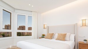 A modern bedroom with a large bed, two bedside tables, a small seating area, and a view of buildings through the windows. - Avenida Andalucía - iloftmalaga (Málaga)