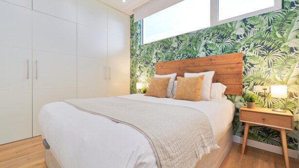 Ein Schlafzimmer mit einem Bett, einem Holzkopfstück, einem Nachttisch und einer Wand mit tropischem Blattmuster.