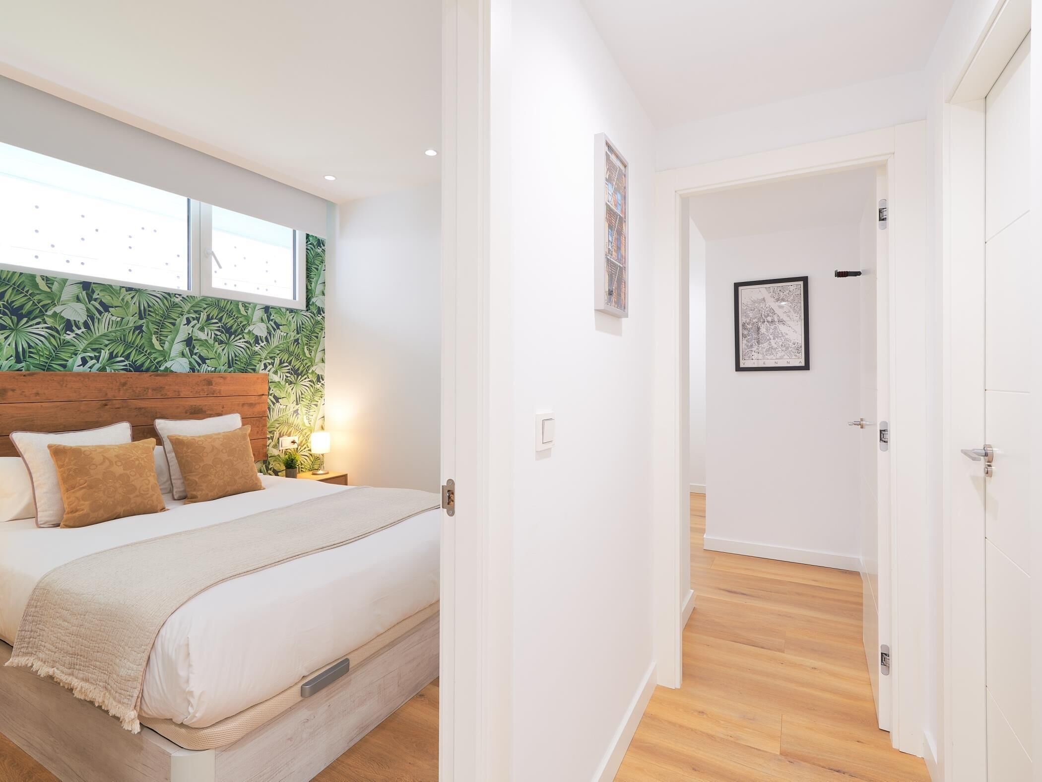 A bedroom with a bed, a nightstand, a wall with a cityscape picture, and a door leading to another room.