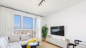 Un salon moderne avec un canapé, une petite table, un téléviseur et une vue sur le paysage urbain grâce à de grandes fenêtres.
