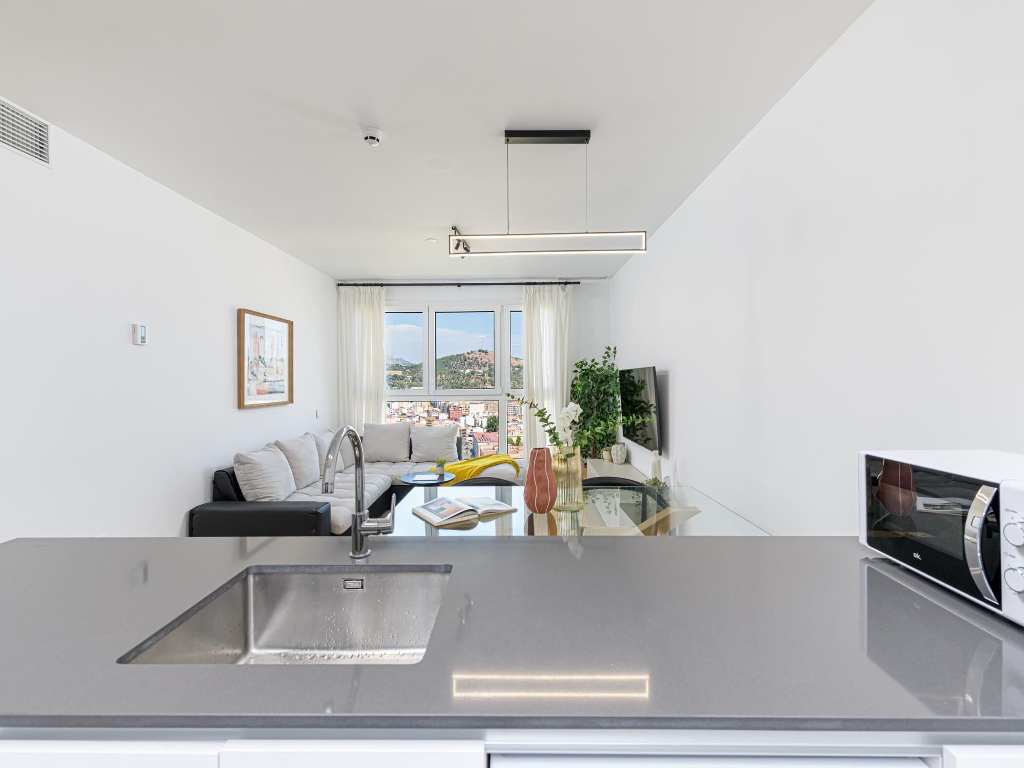 Una cocina moderna con un lavamanos de acero inoxidable, una zona de comedor con sofá y televisor, y vistas a un paisaje urbano a través de amplios ventanales.