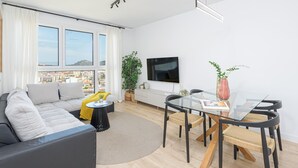 Un salon moderne comprenant un canapé, une table à manger avec des chaises, un téléviseur fixé au mur et une grande fenêtre donnant sur la ville.