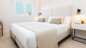 A modern bedroom with two beds, a large window, and a bedside table with a lamp.