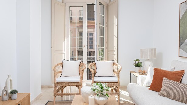 Un salon moderne avec deux fauteuils en osier, un canapé, une table basse et une vue sur un immeuble par une porte ouverte.