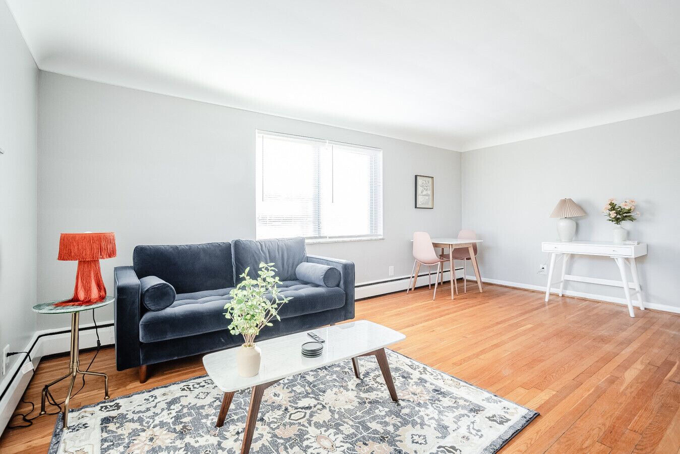 A modern living room with a dark blue sofa, a small coffee table, a plant, a lamp, and a dining area with a white table and chairs.